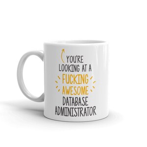 Könnte beinhalten: Weiße Keramiktasse mit Henkel. Die Tasse zeigt den Text "YOU'RE LOOKING AT A FUCKING AWESOME DATABASE ADMINISTRATOR" in schwarzer und gelber Schrift. Die Tasse ist für Datenbankadministratoren konzipiert.