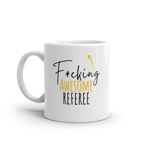 Könnte beinhalten: Weiße Keramik-Kaffeetasse mit einem schwarzen und goldenen Design. Der Text auf der Tasse lautet "F*cking Awesome Referee" mit einem nach oben zeigenden Pfeil.