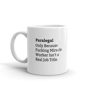 May include: White ceramic coffee mug with a black text design that reads "Paralegal Only Because Fucking Miracle Worker Isn't a Real Job Title."
