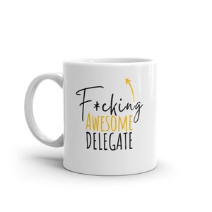 May include: White ceramic coffee mug with a black and gold design. The text on the mug reads "F*cking Awesome Delegate" with an upward pointing arrow.
