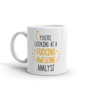 Puede incluir: Taza de café de cerámica blanca con una flecha negra que apunta al texto "You're looking at a fucking awesome analyst" en amarillo.