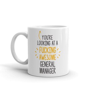 Può includere: Tazza da caffè in ceramica bianca con testo nero che recita "You're looking at a  fucking awesome general manager". Il testo è circondato da trattini gialli. Una freccia punta al testo.