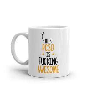 May include: White ceramic mug with a curved handle. The mug features the text "THIS PCSO IS FUCKING AWESOME" in black and orange lettering. The design includes an upward arrow and star accents.