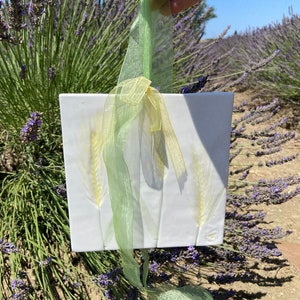 May include: A white ceramic tile with three pressed yellow flower stems. The tile is hanging from a green ribbon.
