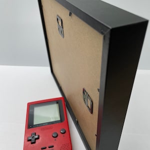 May include: A black picture frame with a cardboard backing and metal hanging hardware. A red handheld video game console is positioned next to the frame. The console has a screen, directional pad, and buttons.