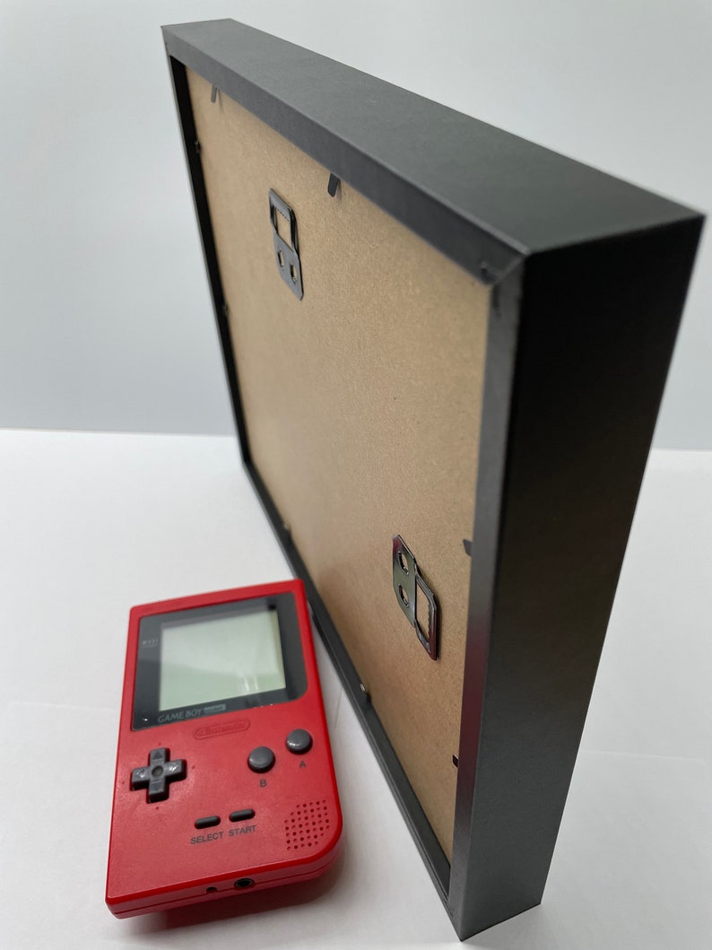 May include: A black picture frame with a cardboard backing and metal hanging hardware. A red handheld video game console is positioned next to the frame. The console has a screen, directional pad, and buttons.