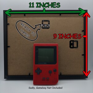 May include: Black picture frame, 28 cm wide and 23 cm tall, with a brown backing. Includes a red Game Boy and the text "READY TO HANG". The text "Sadly, Gameboy Not Included" is also present.