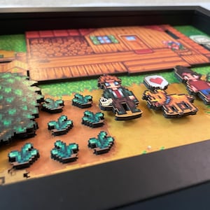 May include: A framed artwork featuring a pixelated farm scene with characters, crops, and a house. The piece includes a brown house, green crops, and characters with a heart symbol. The color scheme is dominated by brown, green, and yellow.