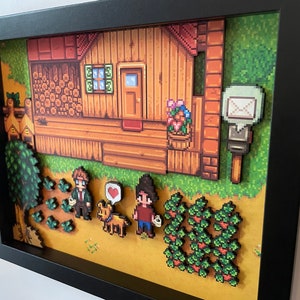 May include: A framed pixel art scene showing a wooden house with a red roof, a mailbox, and characters in a field. The art features a dog with a heart, crops, and a tree. The artwork is in a black frame.