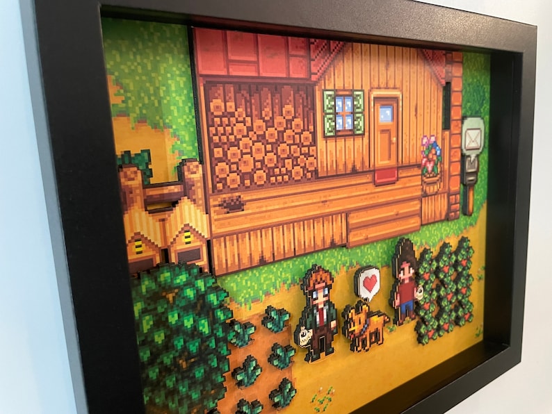 May include: A framed pixel art diorama depicting a wooden house with a door, window, and mailbox. The artwork includes characters, crops, and a dog, all in a retro video game style. The frame is black.