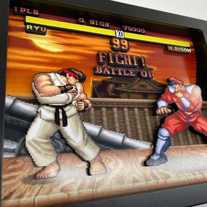 May include: A 3D pixel art diorama of a classic video game fight scene.  The characters Ryu and M. Bison are posed in a fighting stance. The background is a sunset scene with a Japanese temple. The text on the screen reads "KO 99 FIGHT! BATTLE 01".