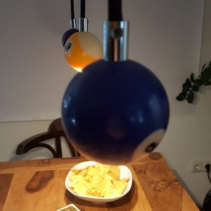 Può includere: Un tavolo di legno con una ciotola di tortilla chips e una piccola ciotola di salsa. Tre lampadari a forma di palla da biliardo pendono dal soffitto sopra il tavolo. Le palle da biliardo sono blu, gialle e viola.