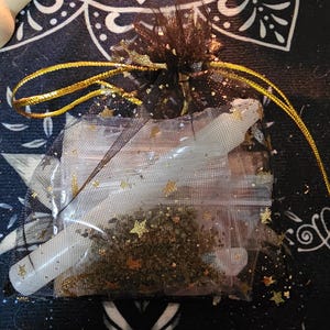 May include: A small, black mesh bag tied with gold ribbon, containing a clear plastic bag filled with herbs and star-shaped glitter. Two white, bone-shaped objects are visible inside the clear bag. The bag sits on a dark blue and black patterned surface.