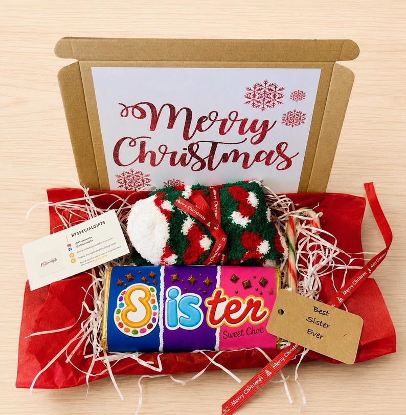 May include: A cardboard gift box containing a pair of green, white, and red fuzzy socks, a chocolate bar with the text Sister Sweet Choc, a candy cane, and a tag reading Best Sister Ever. The box lid features the text Merry Christmas in red.