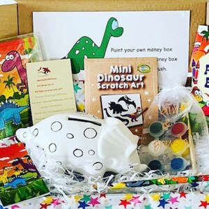 May include: A dinosaur-themed gift box containing a white ceramic piggy bank, paint set, puzzle, scratch art, and a puzzle book. The box includes a card with a green dinosaur illustration and the text "Paint your own money box". The box is filled with colorful tissue paper.
