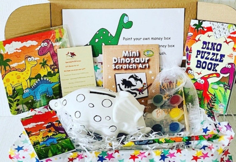 May include: A dinosaur-themed gift box containing a white ceramic piggy bank, paint set, puzzle, scratch art, and a puzzle book. The box includes a card with a green dinosaur illustration and the text "Paint your own money box". The box is filled with colorful tissue paper.