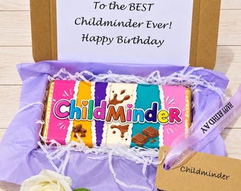 May include: A brown cardboard gift box containing a chocolate bar with a colorful wrapper featuring the text ChildMinder. The box lid has a white card printed with To the BEST Childminder Ever! Happy Birthday. Includes purple tissue paper and a tag.