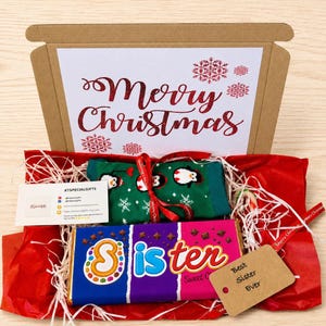 May include: A gift box containing green socks with a penguin pattern, a chocolate bar with a wrapper reading Sister, a white card with Merry Christmas in red glitter text, a small business card, and a brown tag labeled Best Sister Ever.