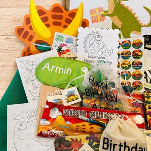 May include: A collection of dinosaur-themed party favors and treats. Includes a dinosaur mask, coloring pages, stickers, a sleep mask with the name "Armin", and a bag labeled "Birthday Treats". The color palette is bright and playful.