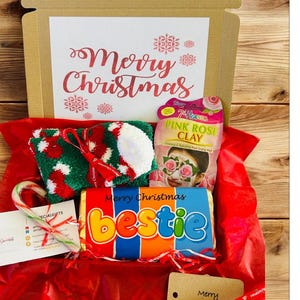 May include: A Christmas gift box filled with festive items. Includes a pair of green, red, and white socks, a candy cane, a face mask, and a chocolate bar that says "bestie". The box is lined with red tissue paper and has "Merry Christmas" on the lid.