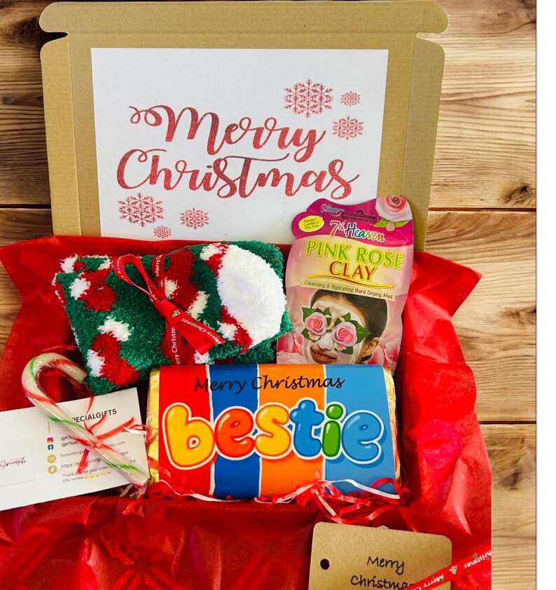 May include: A Christmas gift box filled with festive items. Includes a pair of green, red, and white socks, a candy cane, a face mask, and a chocolate bar that says "bestie". The box is lined with red tissue paper and has "Merry Christmas" on the lid.