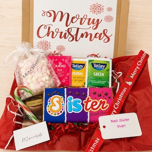 May include: A gift box containing a chocolate bar labeled Sister, a bag of marshmallows, tea bags from Pukka and Tetley, a candy cane, and a hot chocolate packet. The box lid features the text Merry Christmas in red glittery script with snowflake icons.