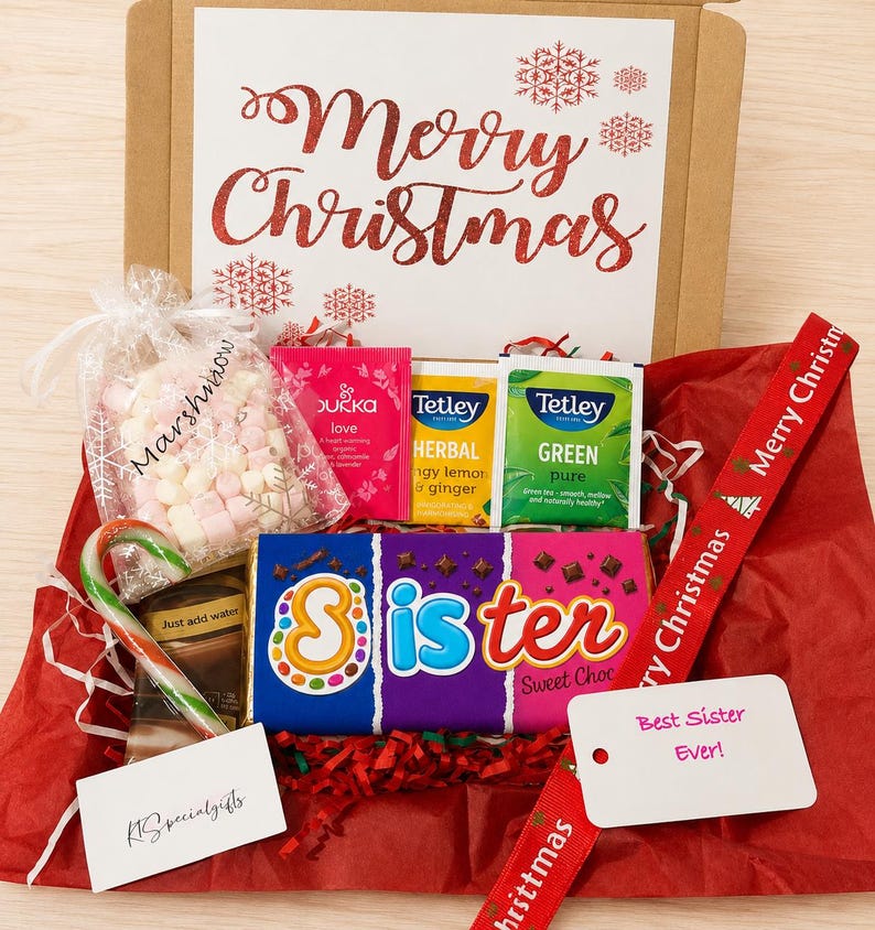 May include: A gift box containing a chocolate bar labeled Sister, a bag of marshmallows, tea bags from Pukka and Tetley, a candy cane, and a hot chocolate packet. The box lid features the text Merry Christmas in red glittery script with snowflake icons.
