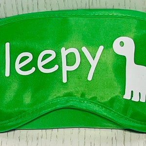 May include: Green eye mask with the word "Sleepy" in white script and a white dinosaur silhouette. The mask is designed to block out light for sleeping. The eye mask is made of a soft, satin-like material.