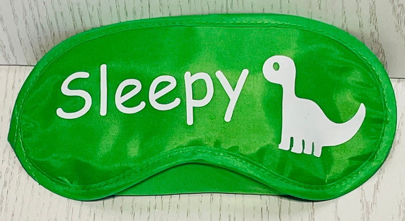 May include: Green eye mask with the word "Sleepy" in white script and a white dinosaur silhouette. The mask is designed to block out light for sleeping. The eye mask is made of a soft, satin-like material.