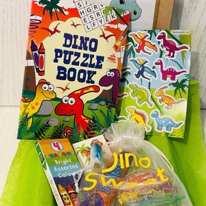 May include: A collection of dinosaur-themed items, including a puzzle book, stickers, crayons, and a bag of sweets. The puzzle book features colorful illustrations and the text "DINO PUZZLE BOOK". The bag is labeled "Dino Sweet".