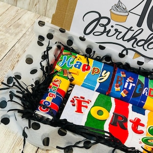 May include: A gift box containing chocolate bars with colorful wrappers featuring the text Happy Birthday and Forty. The box includes black shredded paper filler and white tissue paper with black polka dots. The lid displays a cupcake illustration.