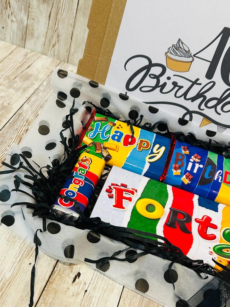 May include: A gift box containing chocolate bars with colorful wrappers featuring the text Happy Birthday and Forty. The box includes black shredded paper filler and white tissue paper with black polka dots. The lid displays a cupcake illustration.