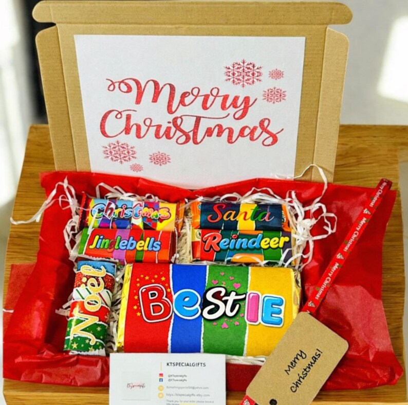 May include: A Christmas gift box with a red lining, filled with colorful candy bars. The box lid features the words "Merry Christmas" in red script. A tag with "Merry Christmas!" hangs from a red ribbon.