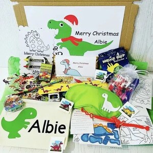 May include: A Christmas-themed gift box filled with dinosaur-themed items. Includes a green dinosaur illustration with the name "Albie", crayons, coloring pages, and a green eye mask. The box also contains the text "Merry Christmas Albie".