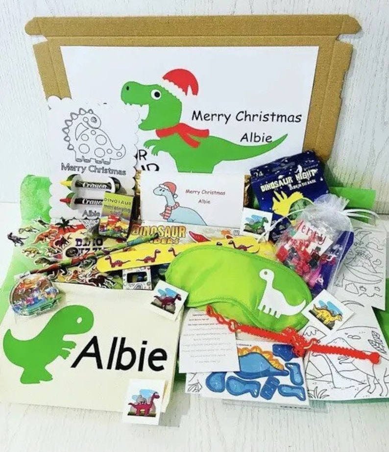May include: A Christmas-themed gift box filled with dinosaur-themed items. Includes a green dinosaur illustration with the name "Albie", crayons, coloring pages, and a green eye mask. The box also contains the text "Merry Christmas Albie".