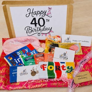 May include: A 40th birthday gift box containing a pink sleep mask, various tea packets, a chocolate bar labeled FORTY, a card with the text Happy 40th Birthday, and a small metal charm, all arranged on pink wrapping paper with gold text.