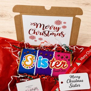 May include: A brown cardboard gift box containing a chocolate bar with a wrapper that reads Sister Sweet Choc. The box lid features the text Merry Christmas in red. Included are red tissue paper, a red ribbon, and a white gift tag reading Merry Christmas Sister.