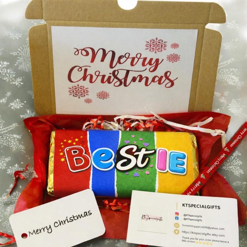 May include: A Christmas gift box with a chocolate bar that says "Bestie" in colorful letters. The box includes a card that says "Merry Christmas" and a red pen with "Merry Christmas" printed on it. The box is brown and lined with red tissue paper.