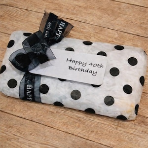 May include: A rectangular gift wrapped in white tissue paper with black polka dots, tied with a black sheer ribbon featuring the text "HAPPY BIRTHDAY" in white, and a white gift tag with the handwritten text "Happy 40th Birthday".