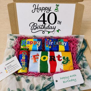 May include: A cardboard gift box containing chocolate bars with colorful wrappers. The box lid features the text Happy 40th Birthday. Inside, the bars are labeled Happy, Birthday, Forty, and Congrats, nestled in pink shredded paper filler.