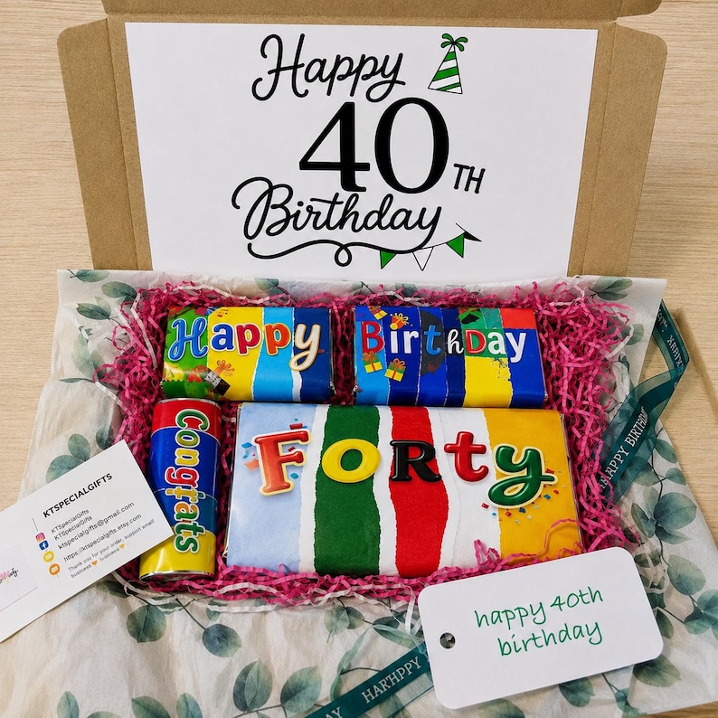 May include: A cardboard gift box containing chocolate bars with colorful wrappers. The box lid features the text Happy 40th Birthday. Inside, the bars are labeled Happy, Birthday, Forty, and Congrats, nestled in pink shredded paper filler.