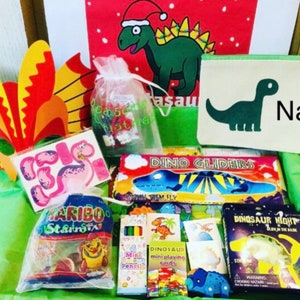 May include: A gift box filled with dinosaur-themed toys and treats. Includes a green pencil case with a dinosaur graphic and the word "Name", a bag of Haribo candies, stickers, and a dinosaur mask. Also includes mini playing cards and a "Dino Gliders" toy.