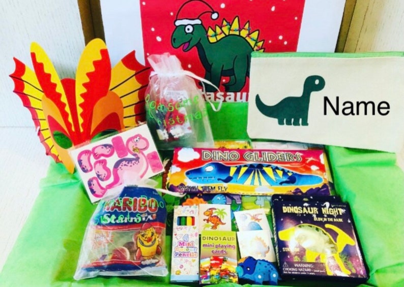 May include: A gift box filled with dinosaur-themed toys and treats. Includes a green pencil case with a dinosaur graphic and the word "Name", a bag of Haribo candies, stickers, and a dinosaur mask. Also includes mini playing cards and a "Dino Gliders" toy.