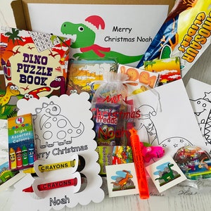 May include: A gift box filled with dinosaur-themed items. Includes a puzzle book, coloring pages, crayons, candy, and a bubble wand. The box is decorated with a dinosaur wearing a Santa hat and the text "Merry Christmas Noah".