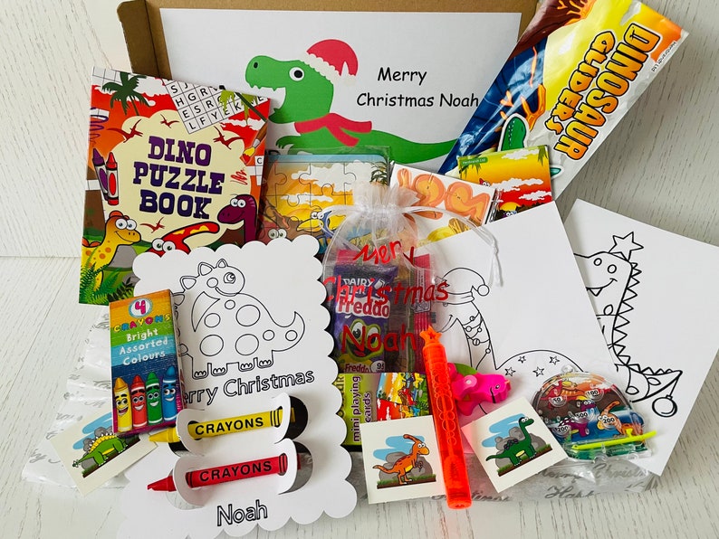 May include: A gift box filled with dinosaur-themed items. Includes a puzzle book, coloring pages, crayons, candy, and a bubble wand. The box is decorated with a dinosaur wearing a Santa hat and the text "Merry Christmas Noah".
