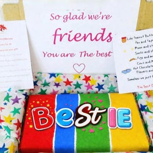 May include: A gift box with a colorful "Bestie" candy bar and handwritten notes. The box contains a candy bar with red, blue, green, and yellow sections. The notes say "So glad we're friends" and list various items like peanut butter and jelly.
