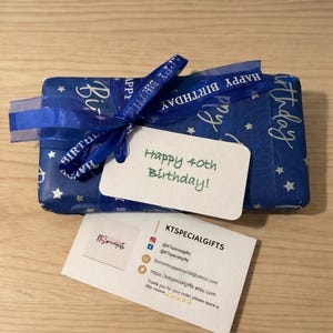 May include: A rectangular gift wrapped in blue paper with white stars and a blue ribbon printed with the words Happy Birthday. A white tag attached to the ribbon reads Happy 40th Birthday! A business card for KTSPECIALGIFTS is placed below the gift.