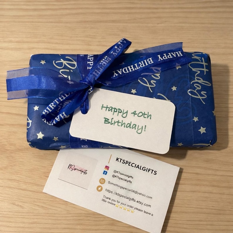 May include: A rectangular gift wrapped in blue paper with white stars and a blue ribbon printed with the words Happy Birthday. A white tag attached to the ribbon reads Happy 40th Birthday! A business card for KTSPECIALGIFTS is placed below the gift.
