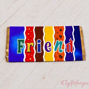 May include: A wrapped chocolate bar with the word "FRIEND" in colorful letters. The packaging has a blue, red, brown, yellow, and purple design with a torn paper effect. The bar is on a light wood surface.