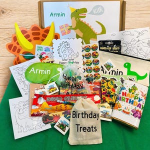 May include: A dinosaur-themed birthday gift set with various items. Includes a cardboard box, a green eye mask, a canvas bag with "Armin" and a dinosaur graphic, stickers, coloring pages, and a birthday-themed cup. The box has a dinosaur illustration and the name "Armin".